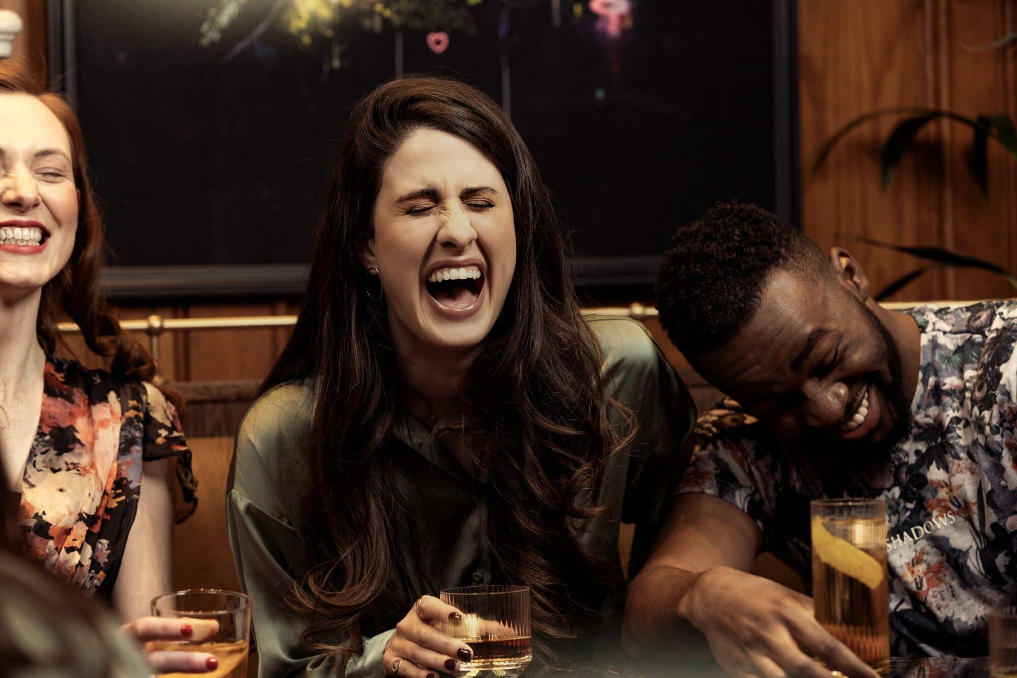 A woman laughing with friends at a dinner table, drinks in hand